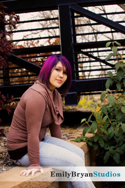Young woman with purple hair sitting next to a bridge.