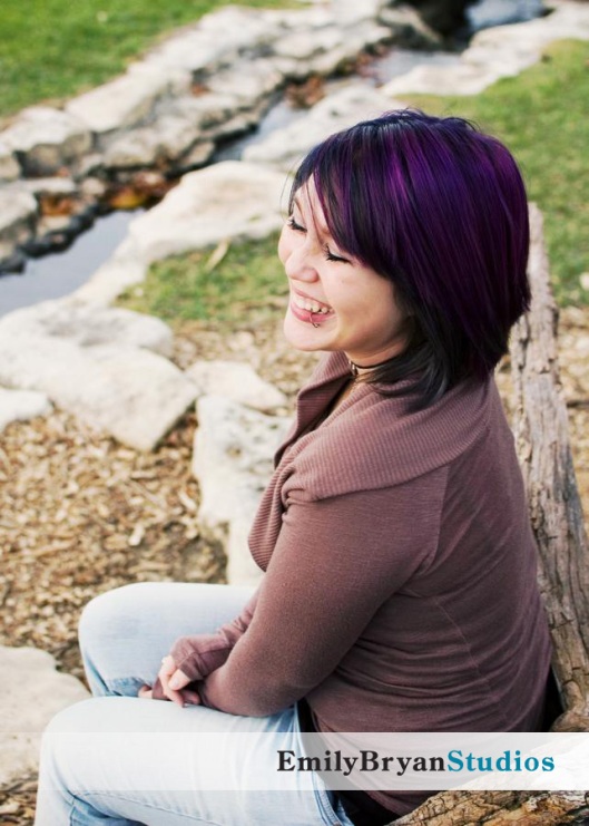 Laughing young woman with purple hair sitting by a stream of water in a park.